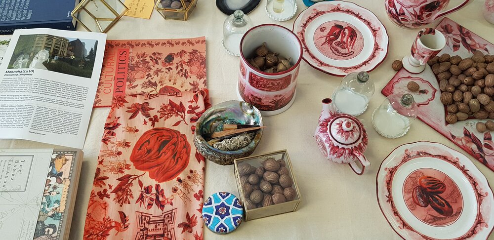 Many items are laid out across a table. There is a newsletter, a history book, a "Cultural Politics" booklet, a red floral tea towel, a bowl containing dried sage, a box of walnuts, and a teapot.