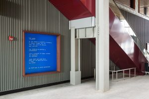 A large vinyl installation of a poem hangs below a staircase. The poem "The News" reads: "in the remote illumination of possibility / a storm cleared the sky / and the crisis of waiting / was relieved // the duration of anything is / unknowable // just a sequence of unreal windows / of contagion / of recovery // shed what was never real / what do you mean have faith / i love / to know".