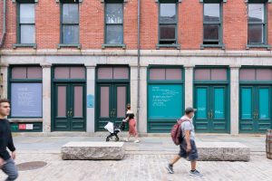 People walk by large window installations of poems on a New York City street. A woman with a stroller pauses to look at the installation.