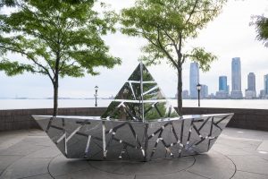 Metal sculpture of a boat with a background of water and trees and buildings