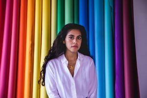 A woman, journalist and artist Mona Chalabi, poses in front of a colorful rainbow background