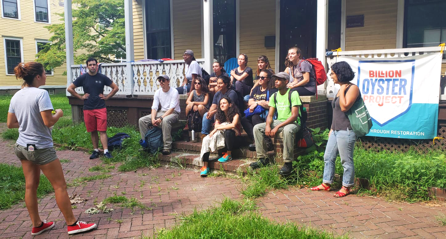 A group of people sit on a stoop, in front of a Billion Oyster Project banner, at a historic house in Nolan Park on Governors Island.