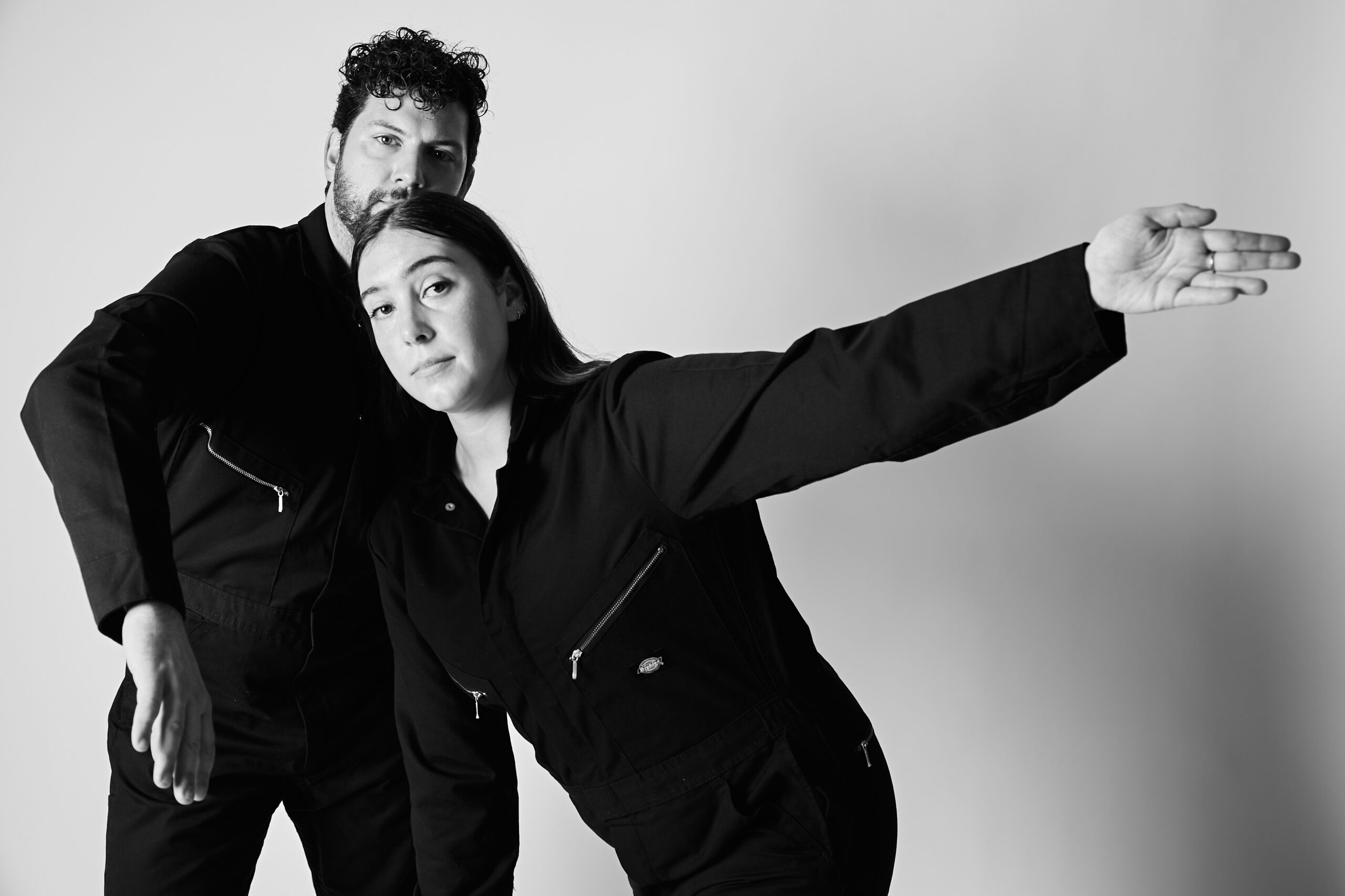 Black and white photo of two overlapping bodies in black jumpsuits against a white background. Nattie is leaning forward with long dark hair and one arm extended in a long line to the side, Hollis is behind her more upright with curly dark hair and one arm bent forward.