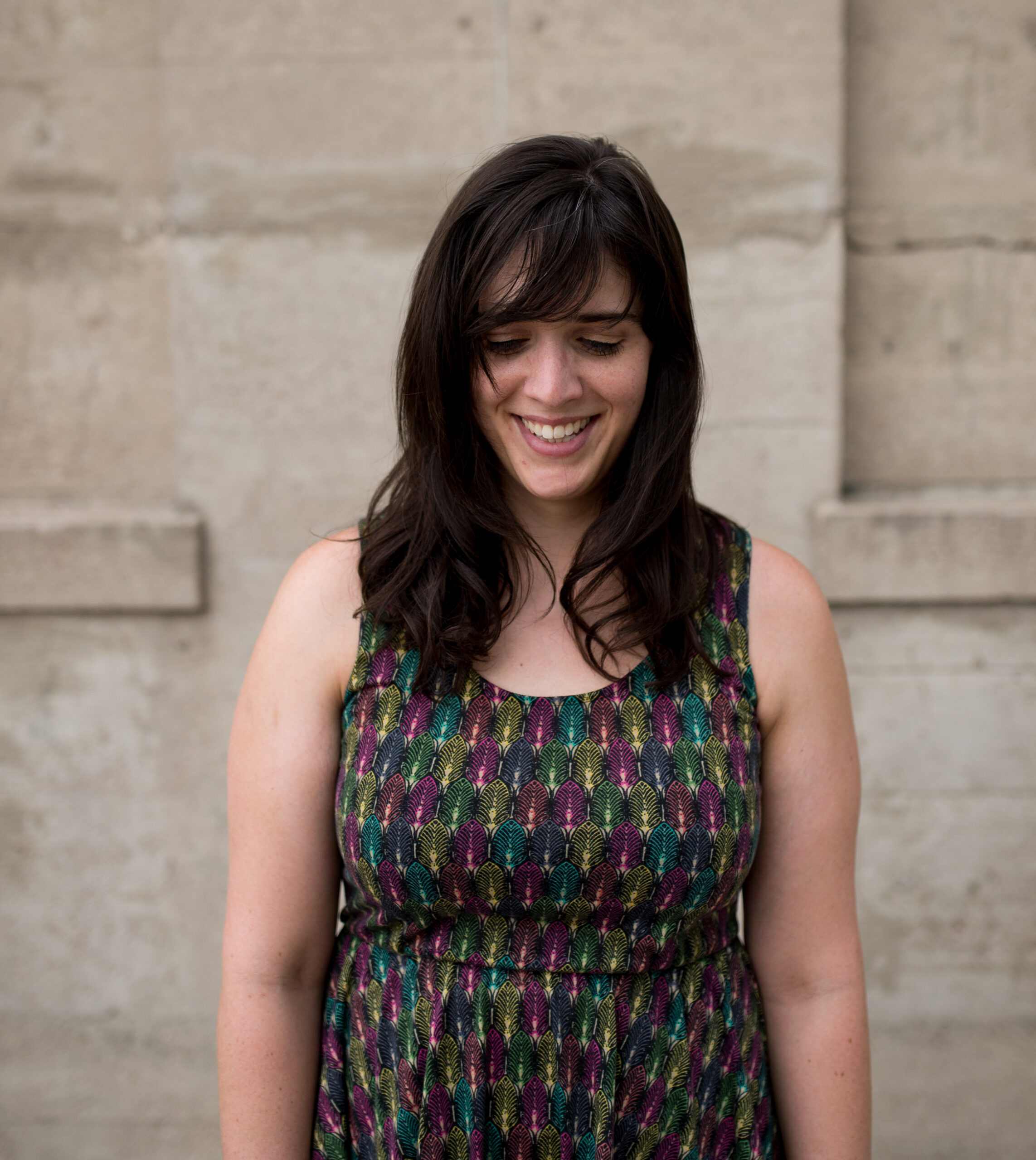 This is a headshot of Kate Speer, who is a white woman. She is standing in front of a blurred concrete wall. She is wearing a sleeveless dress with a multi-colored pattern. She is smiling with her gaze looking down. She has shoulder length dark brown hair and bangs.