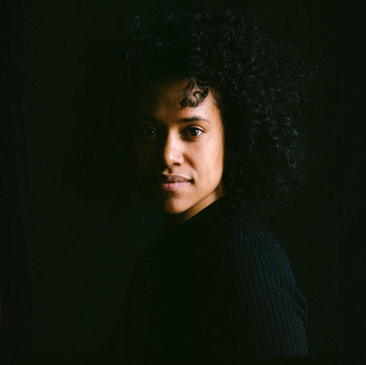 Portrait of a Black woman with dark, curly hair and wearing a black sweater staring straight at the camera, with the right side of her face obscured by shadow.