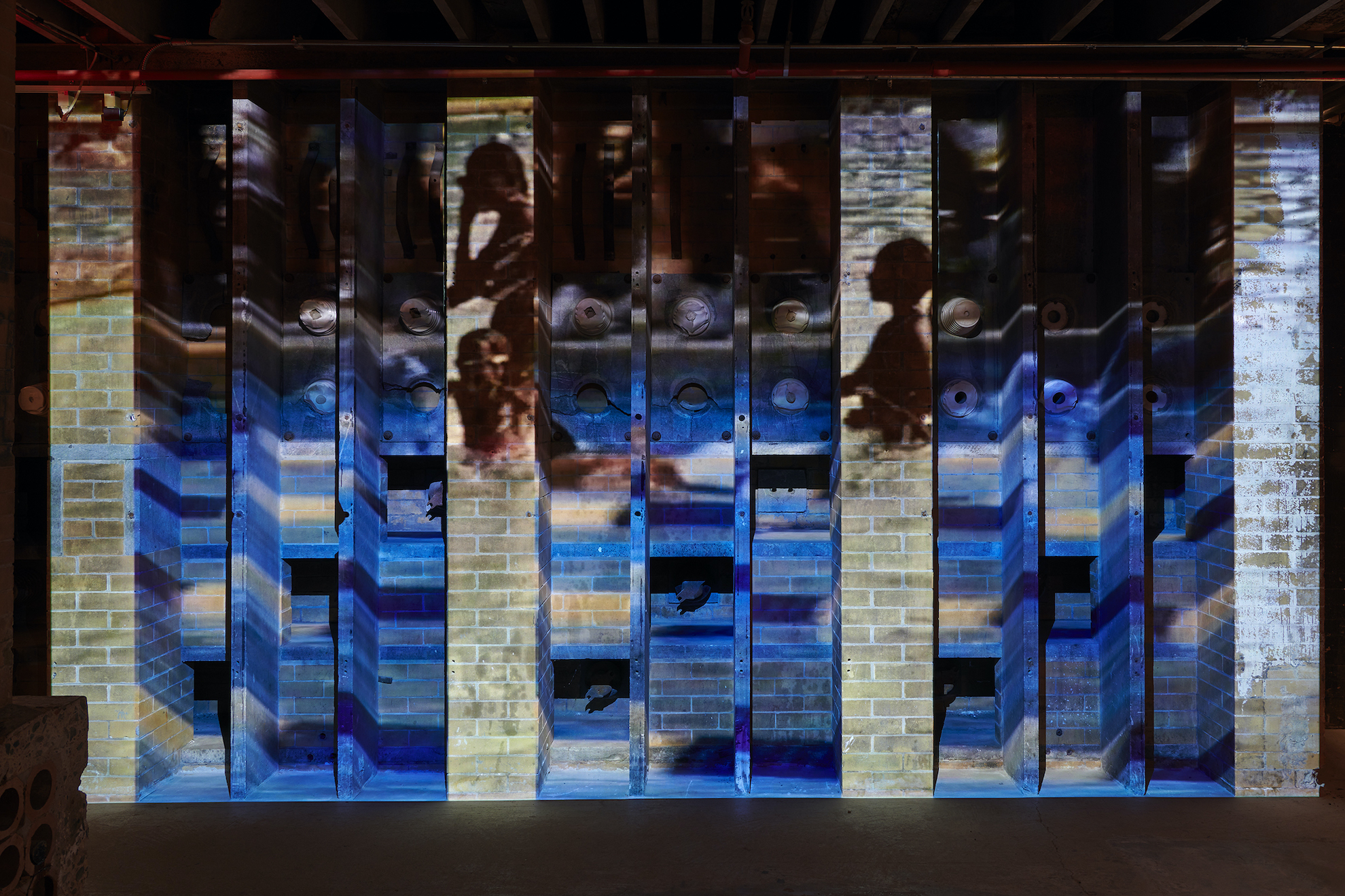 A video projection onto a series of recessed brick walls illuminates a Black woman and children in a swimming pool amidst blue waves.