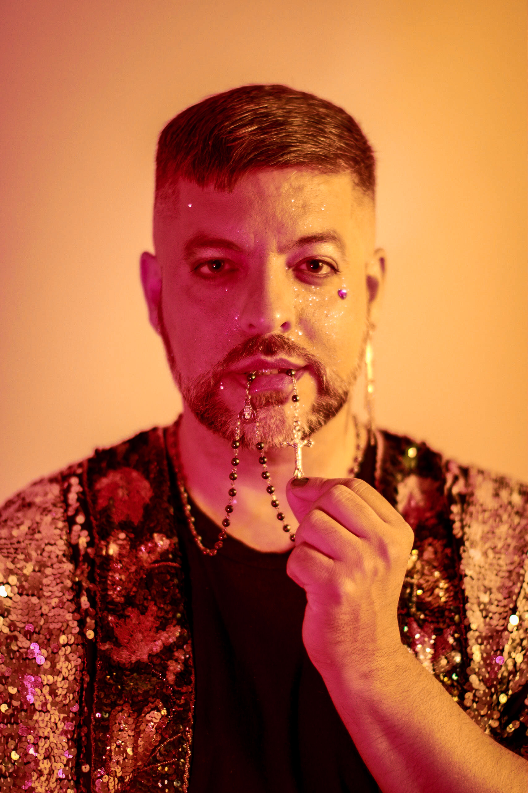 A cis, Latinx man with short dark hair and a goatee beard fingers the cross on a rosary, suspended from his mouth. He is wearing a sequined jacket over a black shirt, and photographed against a glowing orange background.