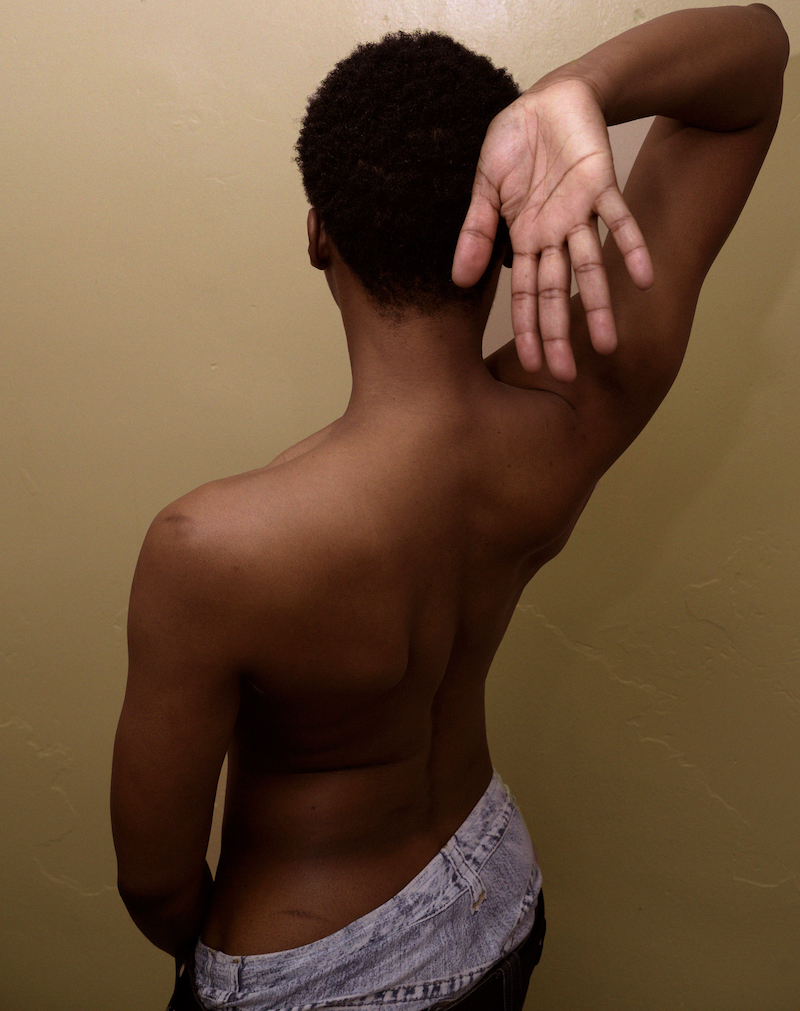 NIC Kay, a gender non-conforming (GNC) individual of Black descent, is prominently featured in this image. Their shortly cropped natural hair frames their face elegantly. NIC Kay’s bare back and right arm extend gracefully over the right shoulder, with the right hand wide and facing towards the camera. They are clad in oversized denim jeans, exuding a sense of casual confidence.