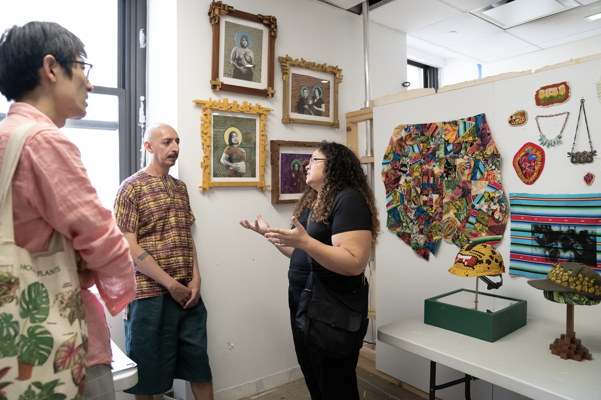 Three individuals standing in an artist studio having a conversation. Behind the individuals are two corner studio walls filled with colorful artwork including framed photo portraits, textile pieces, jewelry and sculptures of construction helmets, displayed on a table.