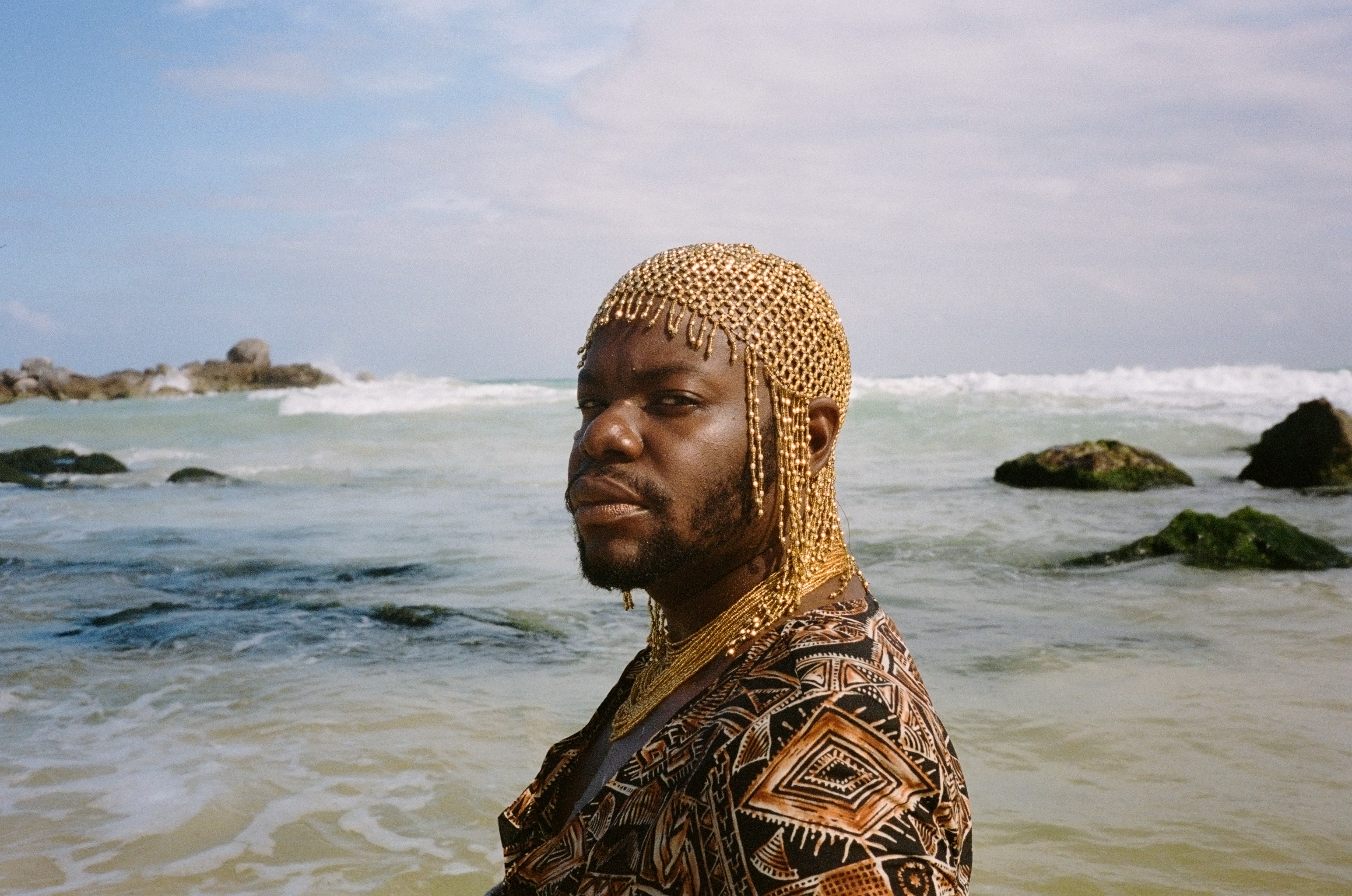 jaamil olawale kosoko stands in front of a light blue body of water, looking into the camera. jaamil is wearing a top with gold and black print and a beaded gold headpiece.