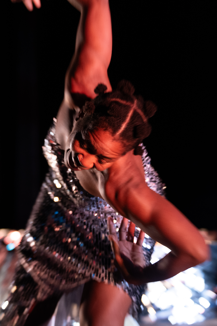 mayfield brooks, a chestnut brown skinned individual, wears sparkly dress moving toward the camera with the arms extended like wing. The background is black and the image is vertical.