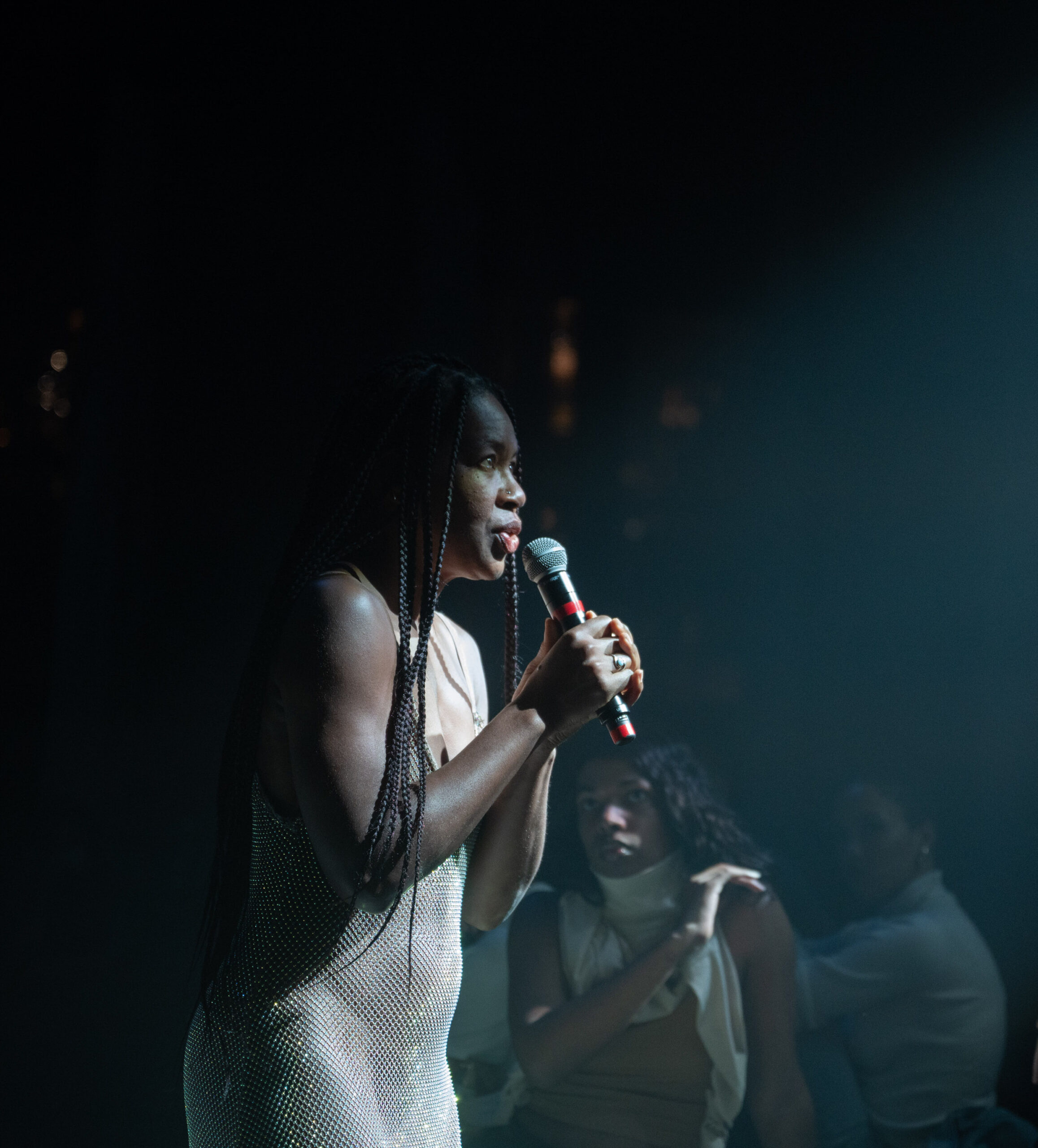 mayfield brooks, a chestnut brown skinned individual, is wearing a sparkly dress and visible in the glow of a single spotlight that illuminates them holding a microphone and speaking to seating audience members that surround them.