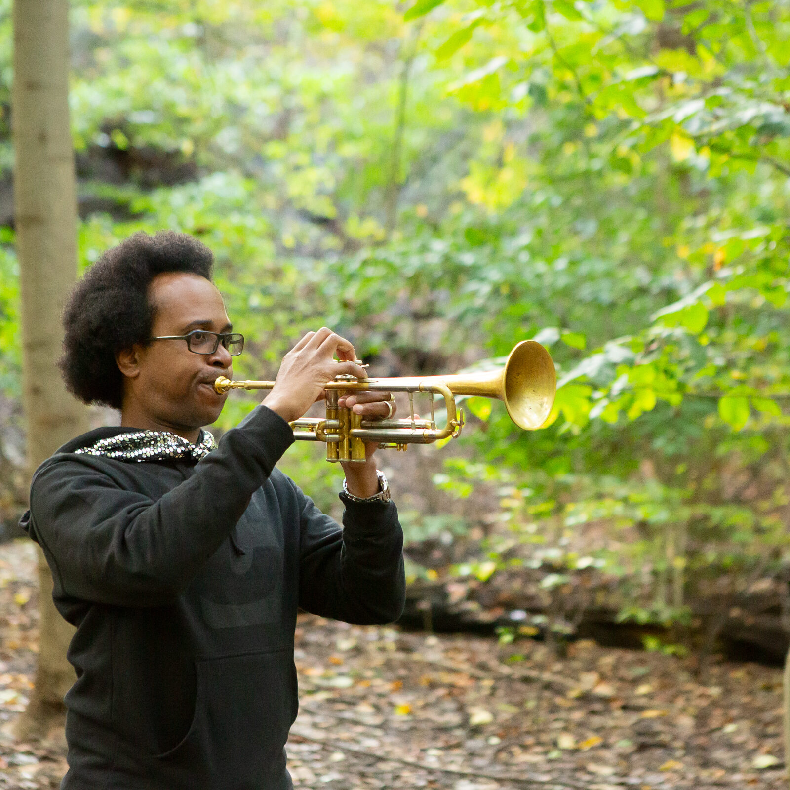 In this image, a person wearing black clothing is playing the trumpet. They are outside with green leaves and many trees in the background.