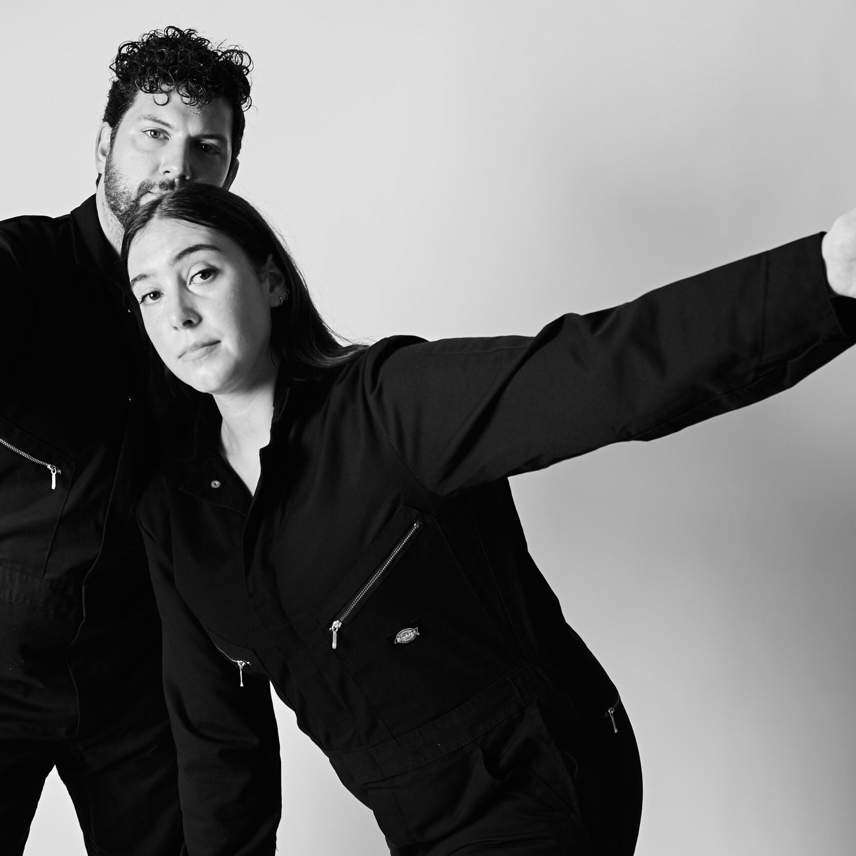 Black and white photo of two overlapping bodies in black jumpsuits against a white background. Nattie is leaning forward with long dark hair and one arm extended in a long line to the side, Hollis is behind her more upright with curly dark hair and one arm bent forward.