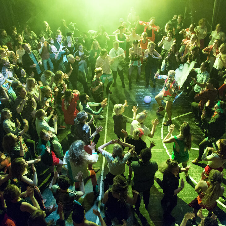 A large group of people in a gymnasium surround the artist, dressed in a costume evoking bubbles and high-energy aerobics, who guides them in movement. The participants are bathed in a green-glowing light and photographed from above.