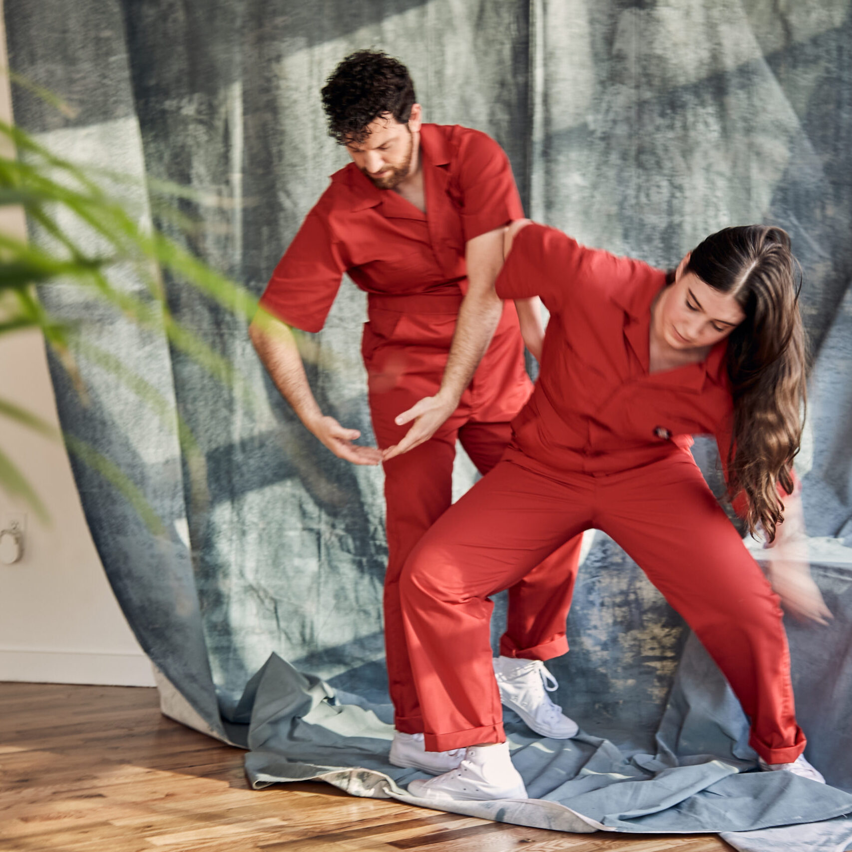 Two dancers in red jumpsuits are caught mid movement in front of a gray drape, a few green plant fronds poke into the foreground of the frame.