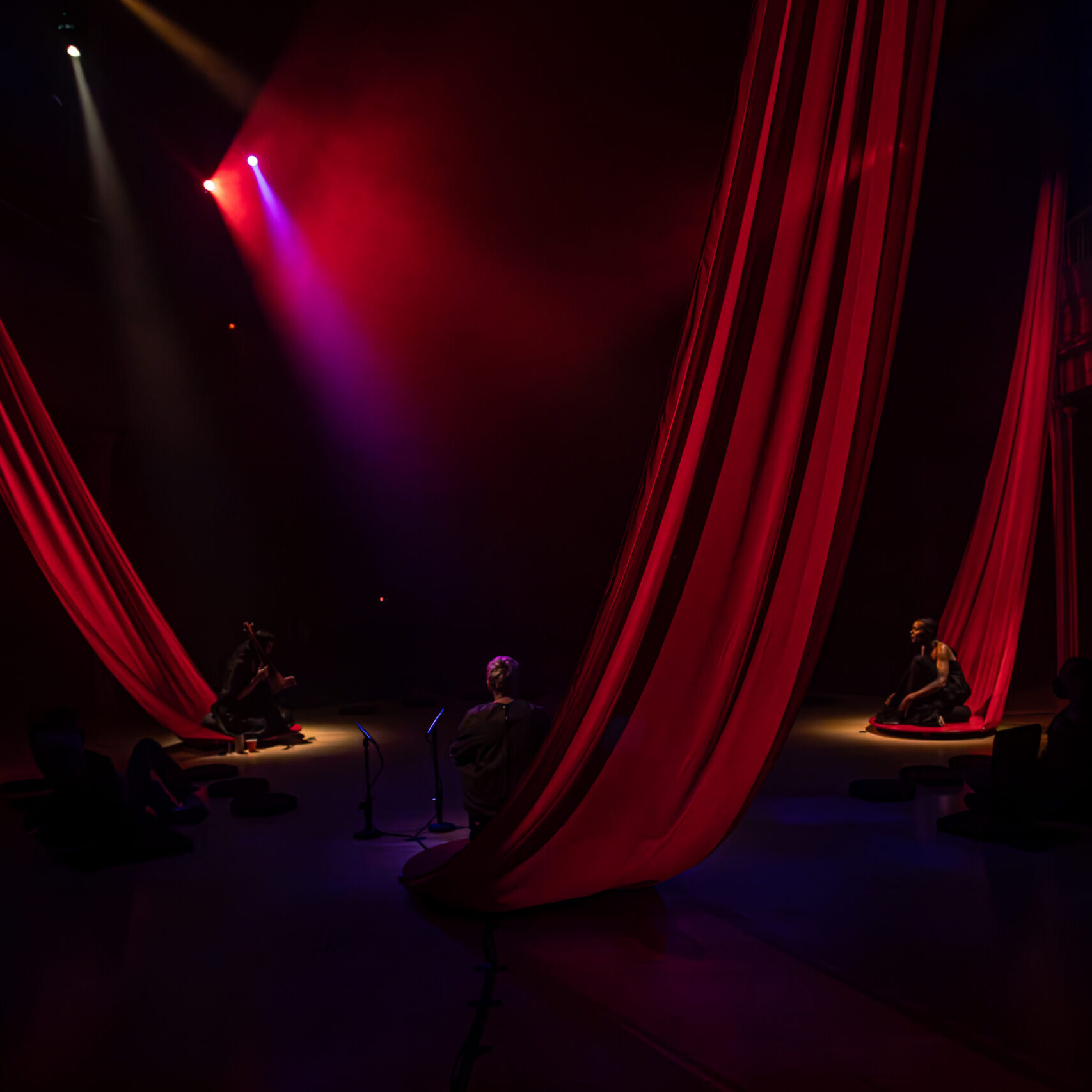 Three large red drapes are hung from the ceiling to the floor in the shape of a triangle. Three people are sitting on each piece of fabric on the floor. The room is dark with two high bright lights shining down in the upper left corner.