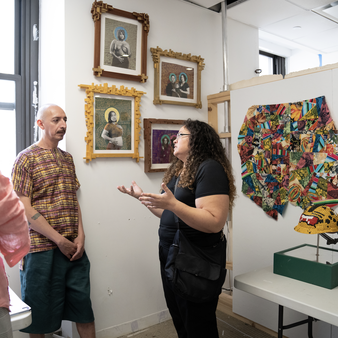 Three individuals standing in an artist studio having a conversation. Behind the individuals are two corner studio walls filled with colorful artwork including framed photo portraits, textile pieces, jewelry and sculptures of construction helmets, displayed on a table.