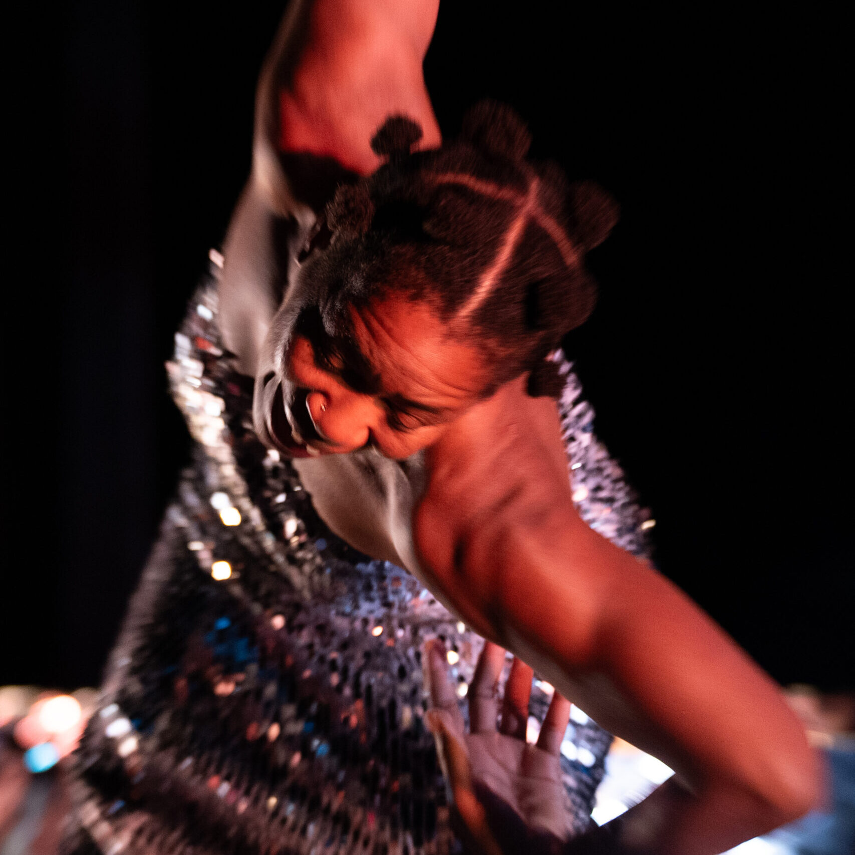 mayfield brooks, a chestnut brown skinned individual, wears sparkly dress moving toward the camera with the arms extended like wing. The background is black and the image is vertical.