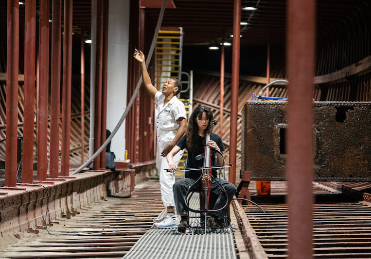 mayfield brooks is depicted in the hold of the Wavertree ship with Dorothy Carlos. mayfield, a chestnut brown skinned individual, is wearing a white boiler suit and turns to the side to vocalize, their mouth open and right arm raised in exclamation. Dorothy, a tan skinned individual wearing a black t-shirt and black pants, is seated and playing an electric cello. – with the spine exposed.