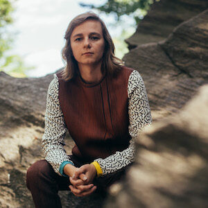 Image of Shannon Finnegan: Me, a white person with shoulder-length, blonde hair looking directly at the camera. I'm wearing my favorite outfit — a wood grain sweatshirt and pant set. The sweatshirt has contrasting sleeves with a squiggle pattern and sleeve cuffs in two different colors. There are rocks and foliage out-of-focus in the background.
